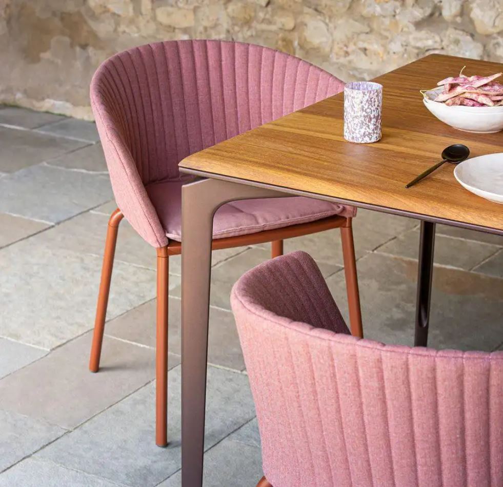 Dos sillas tapizadas de rosa se colocan alrededor de una mesa de madera con un cuenco, un candelabro y una cuchara sobre un suelo de baldosas.